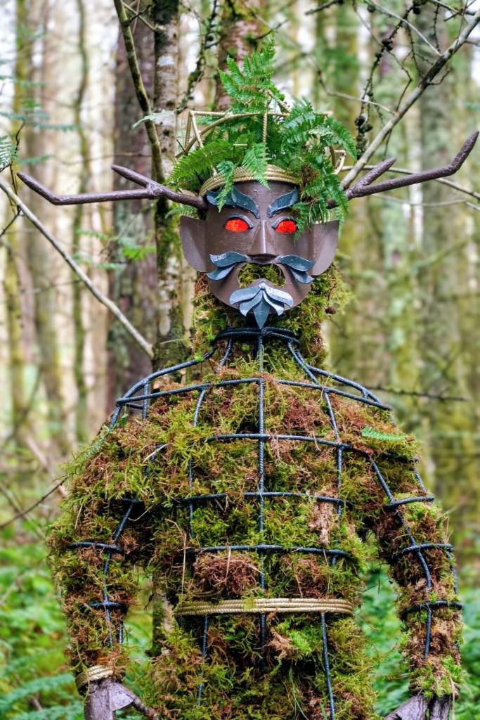 A Green Man sculpture made of moss on a wire frame, with a horned mask, fern crown and red eyes, standing among trees.