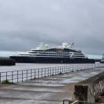 A dark-hulled luxury cruise ship named Le Champlain being guided into Swansea docks by a tug, with a person watching from the waterfront railings under a grey sky.