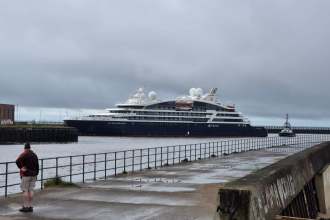 A dark-hulled luxury cruise ship named Le Champlain being guided into Swansea docks by a tug, with a person watching from the waterfront railings under a grey sky.