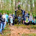 A large group of adults and children gathered around a tall moss-covered Green Man sculpture in woodland, some children climbing on it.