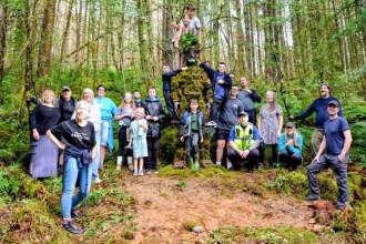 A large group of adults and children gathered around a tall moss-covered Green Man sculpture in woodland, some children climbing on it.