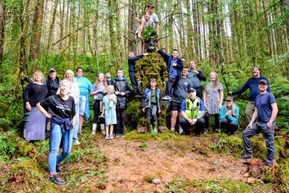 A large group of adults and children gathered around a tall moss-covered Green Man sculpture in woodland, some children climbing on it.