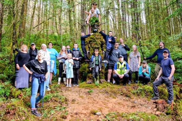 A large group of adults and children gathered around a tall moss-covered Green Man sculpture in woodland, some children climbing on it.