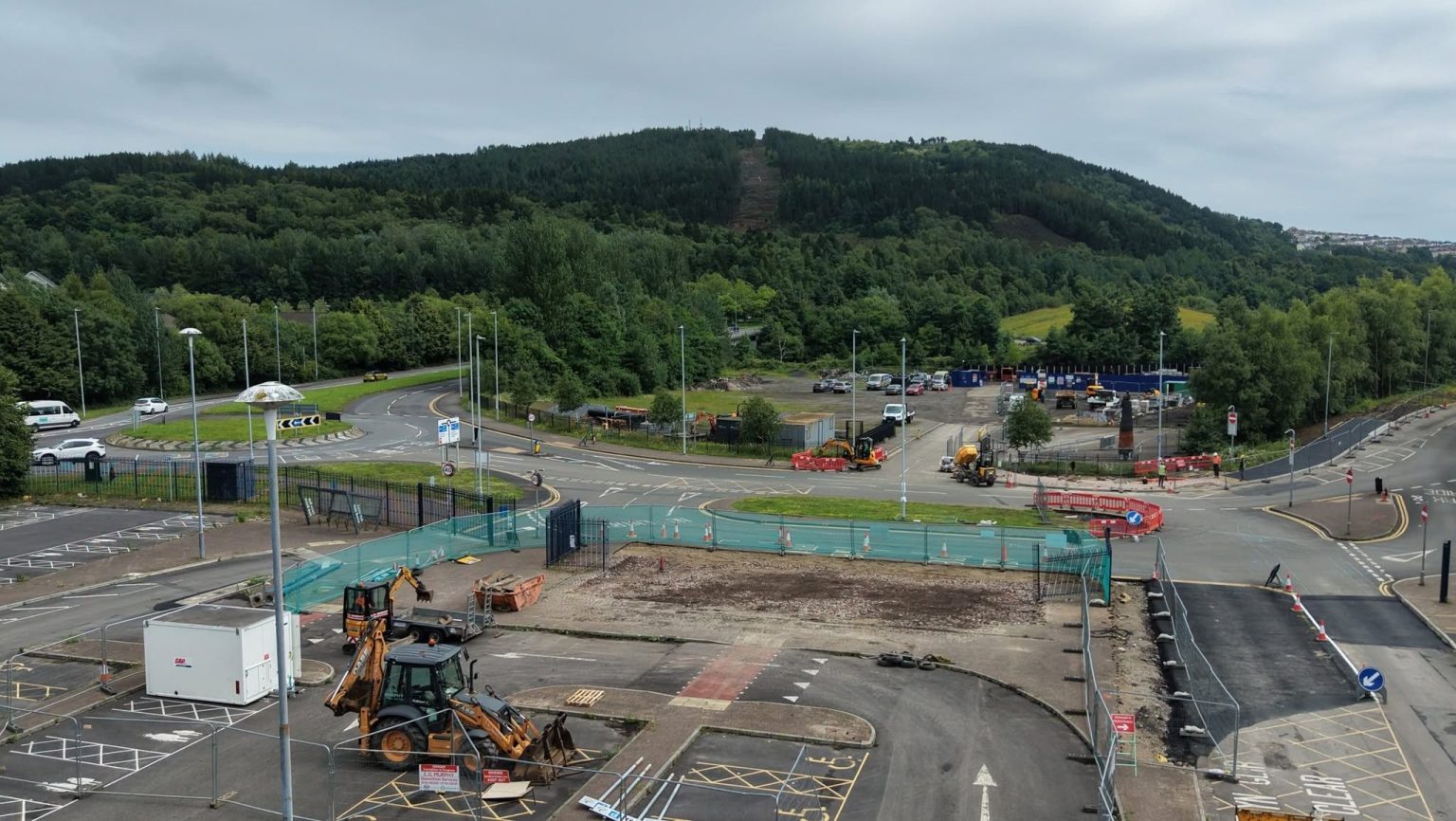 Diggers and contractor cabins on a fenced-off section of Landore Park & Ride car park, with the tree-covered slope of Kilvey Hill rising behind.