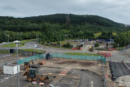 Diggers and contractor cabins on a fenced-off section of Landore Park & Ride car park, with the tree-covered slope of Kilvey Hill rising behind.