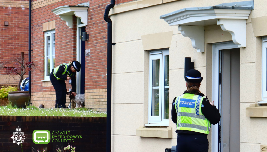 Two police community support officers in high-visibility jackets calling at the front doors of houses on a residential street, one greeting a small dog.