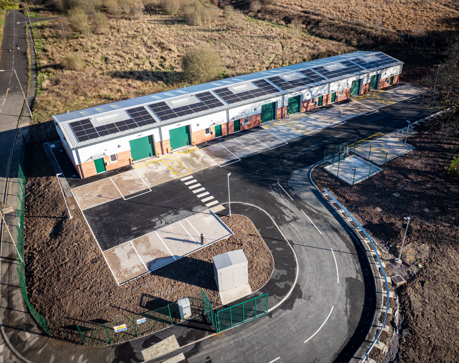 Aerial view of a long single-storey industrial unit with solar panels on its roof and green roller doors, beside an empty car park at Nant y Cafn Business Park, Seven Sisters.