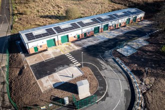 Aerial view of a long single-storey industrial unit with solar panels on its roof and green roller doors, beside an empty car park at Nant y Cafn Business Park, Seven Sisters.
