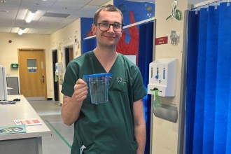 A man in green hospital scrubs smiling and holding a clear plastic jug of water in a hospital emergency department.