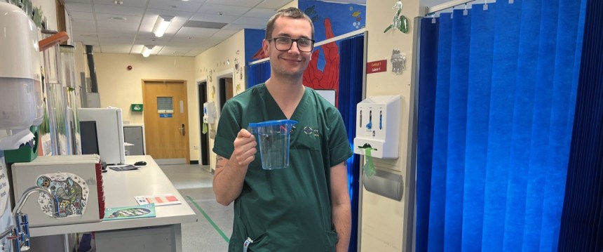 A man in green hospital scrubs smiling and holding a clear plastic jug of water in a hospital emergency department.