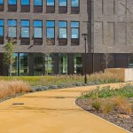 A modern brick health centre building at Canolfan Pentre Awel, with a curved yellow path, bench and landscaped planting in front.
