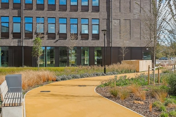 A modern brick health centre building at Canolfan Pentre Awel, with a curved yellow path, bench and landscaped planting in front.