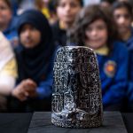 A dark carved Egyptian cippus showing a deity standing over crocodiles, covered in hieroglyphs, on display with schoolchildren out of focus behind.