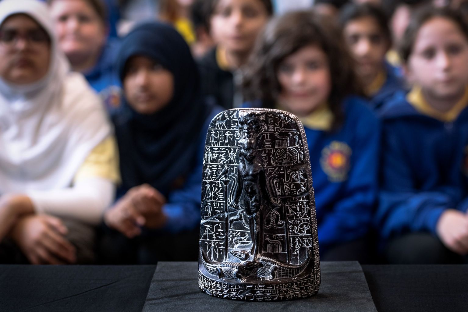 A dark carved Egyptian cippus showing a deity standing over crocodiles, covered in hieroglyphs, on display with schoolchildren out of focus behind.