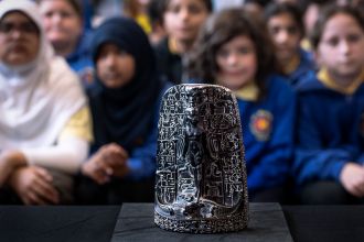 A dark carved Egyptian cippus showing a deity standing over crocodiles, covered in hieroglyphs, on display with schoolchildren out of focus behind.