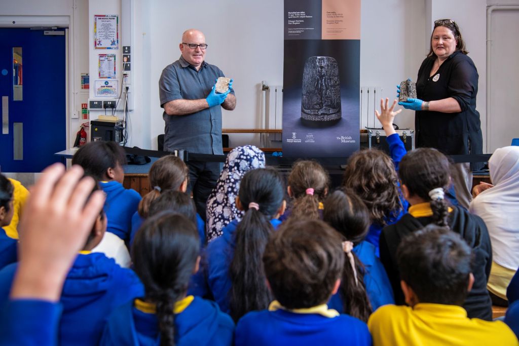Two adults wearing gloves hold ancient artefacts in front of a seated group of schoolchildren, beside an exhibition banner.