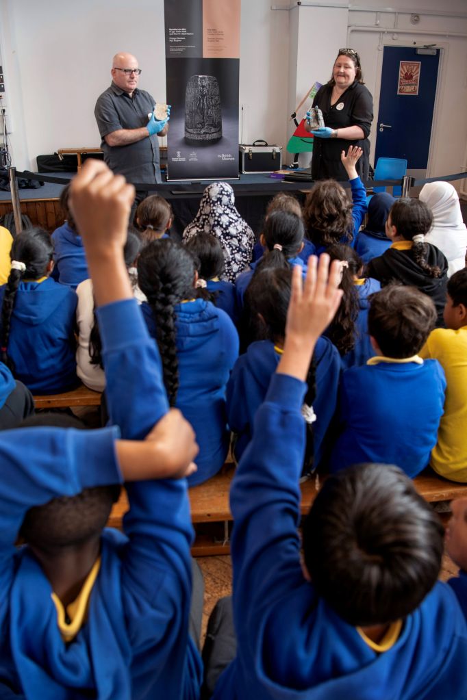 Two adults wearing gloves hold ancient artefacts on a low stage in front of a seated group of schoolchildren with hands raised.