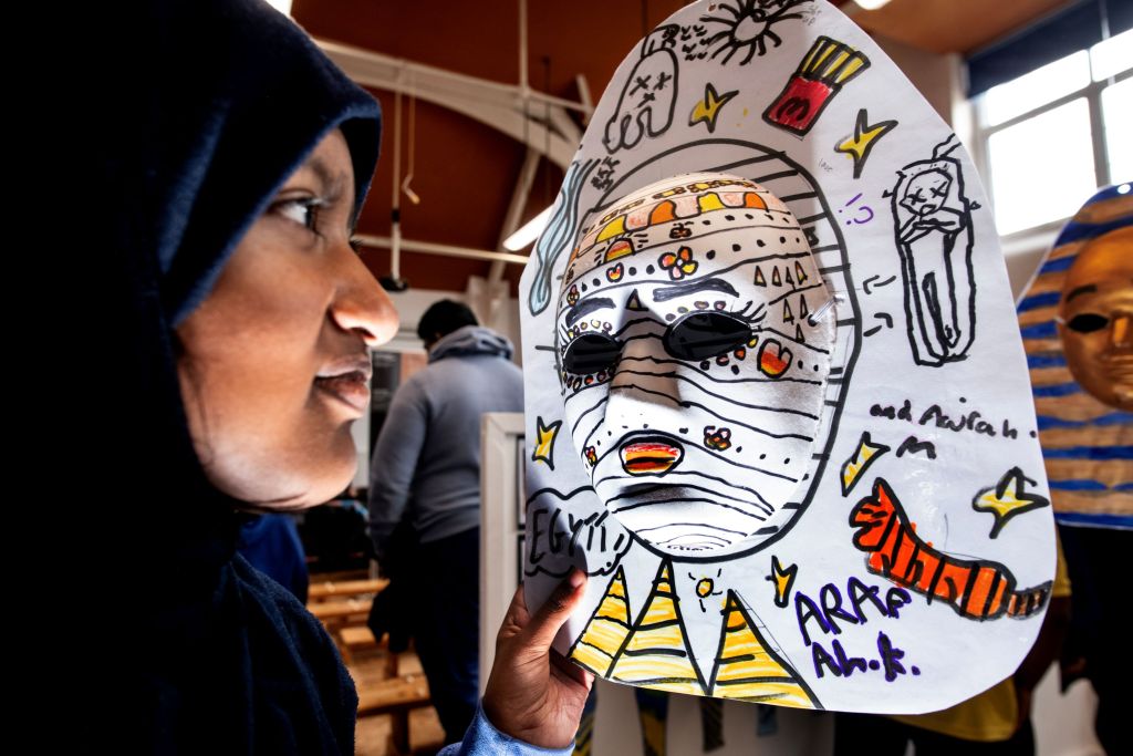 A child in profile holds up a hand-drawn paper mask decorated with an Egyptian mummy design and the word "Egypt".
