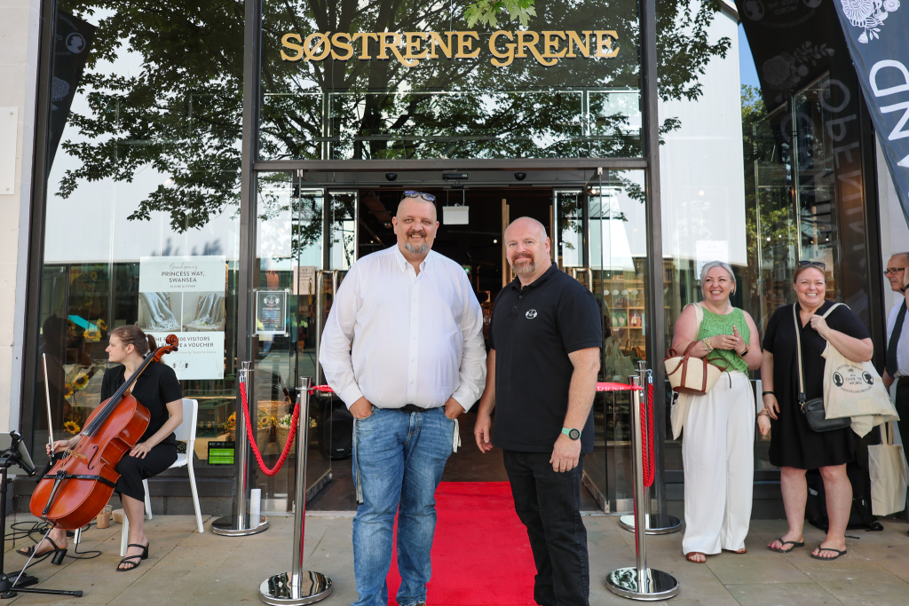 Two men stand smiling on the red carpet at the store entrance under the gold Søstrene Grene sign, with a cellist and well-wishers nearby.