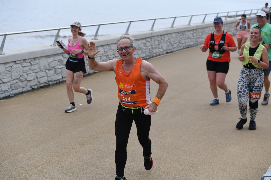 A runner in a Maggie's vest waving while running along Swansea seafront with the sea behind.