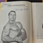 A black-and-white bodybuilding magazine photo of a young Arnold Schwarzenegger, signed in blue ink.
