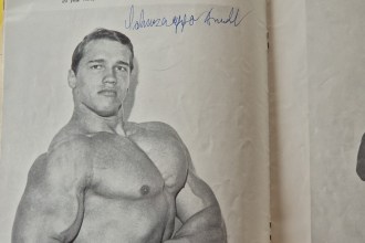A black-and-white bodybuilding magazine photo of a young Arnold Schwarzenegger, signed in blue ink.