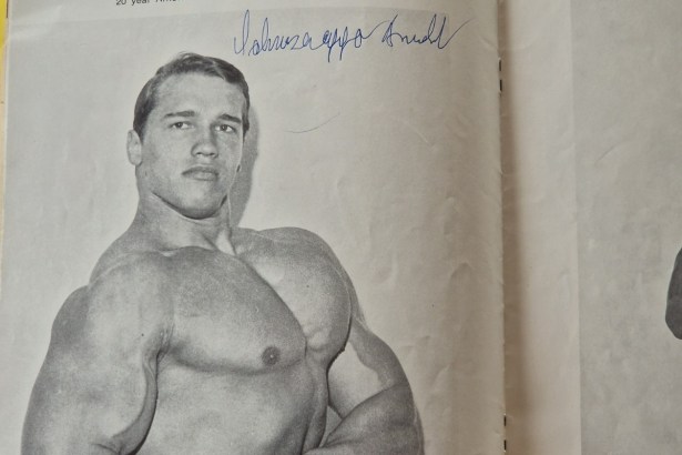 A black-and-white bodybuilding magazine photo of a young Arnold Schwarzenegger, signed in blue ink.