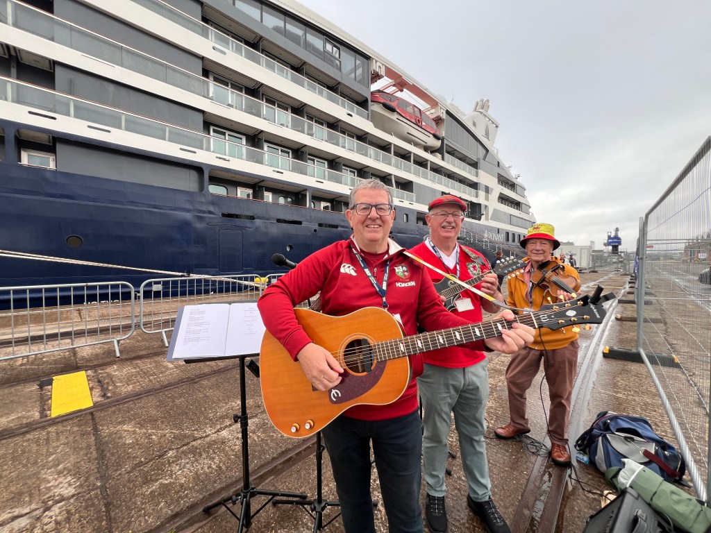 Three musicians with a guitar, mandolin and fiddle performing on a dockside in front of a cruise ship, two wearing Wales rugby shirts.
