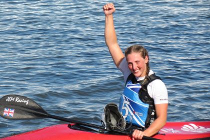 A woman with brown hair platted into pigtails sitting in a red canoe with her arm raised in celebration.