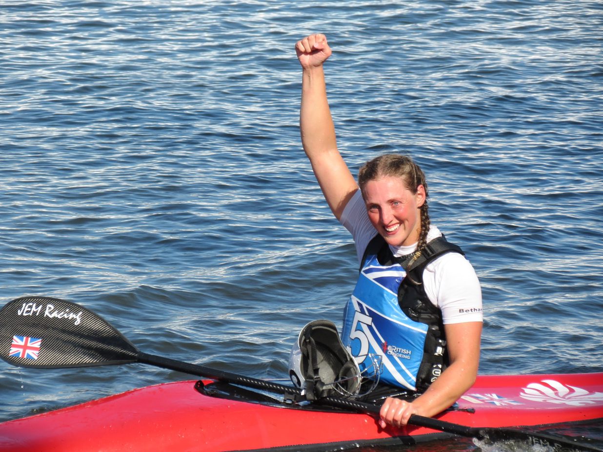 A woman with brown hair platted into pigtails sitting in a red canoe with her arm raised in celebration.