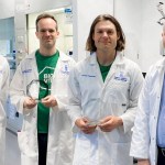 Four men in white lab coats stand in a Swansea University laboratory, two holding glass award trophies.