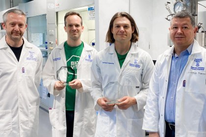 Four men in white lab coats stand in a Swansea University laboratory, two holding glass award trophies.