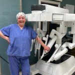 Anita Steel in blue scrubs smiling beside a robotic surgery machine in a hospital theatre.