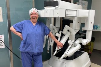 Anita Steel in blue scrubs smiling beside a robotic surgery machine in a hospital theatre.