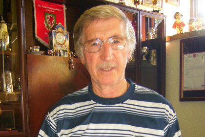 Cliff Jones, silver-haired and wearing a Tottenham Hotspur shirt, holding a book about the 1961 Spurs Double at home surrounded by football trophies and memorabilia