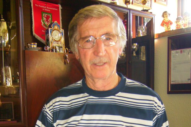 Cliff Jones, silver-haired and wearing a Tottenham Hotspur shirt, holding a book about the 1961 Spurs Double at home surrounded by football trophies and memorabilia