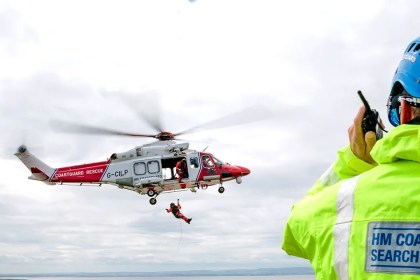 An HM Coastguard search and rescue helicopter winches a casualty from the air as a coastguard officer in a hi-vis jacket radios from a clifftop.
