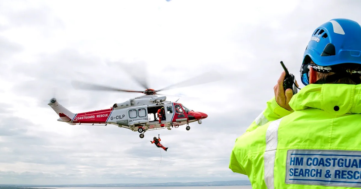 An HM Coastguard search and rescue helicopter winches a casualty from the air as a coastguard officer in a hi-vis jacket radios from a clifftop.