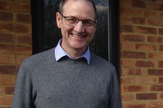 Dr Martin Rolles smiling, wearing glasses and a grey jumper, standing in front of a brick wall.