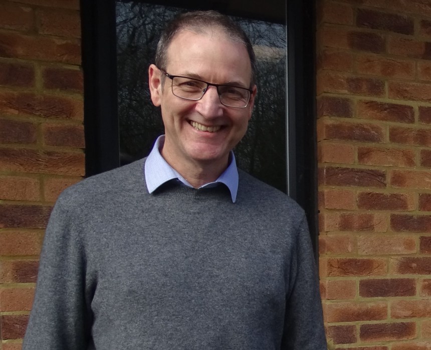 Dr Martin Rolles smiling, wearing glasses and a grey jumper, standing in front of a brick wall.