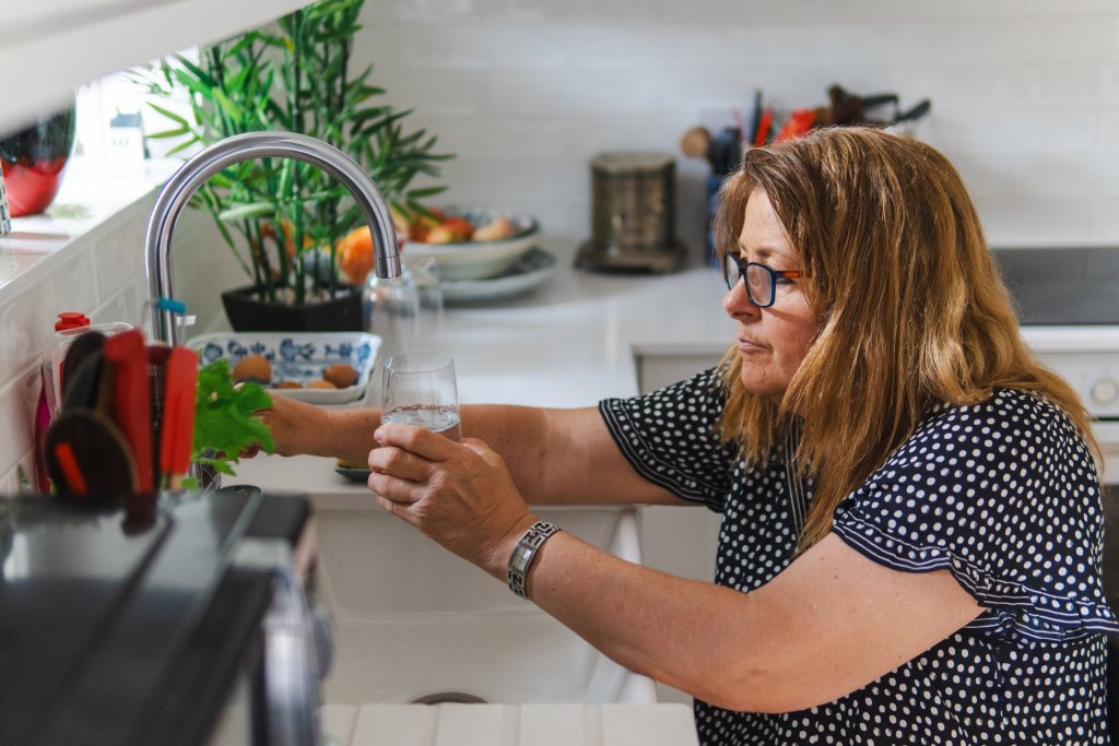 An older woman in a polka-dot top fills a glass of water at her kitchen tap.