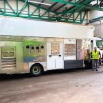 A large white and silver electric recycling vehicle inside a depot, with three council staff in hi-vis standing beside it.
