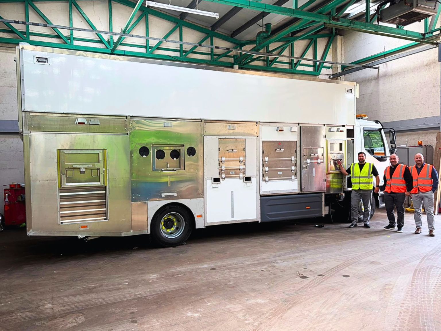 A large white and silver electric recycling vehicle inside a depot, with three council staff in hi-vis standing beside it.