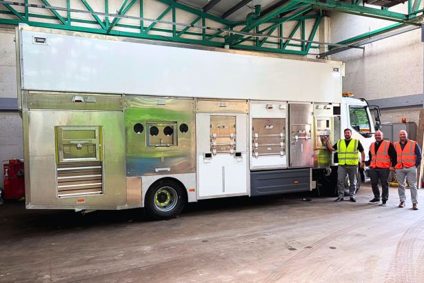 A large white and silver electric recycling vehicle inside a depot, with three council staff in hi-vis standing beside it.