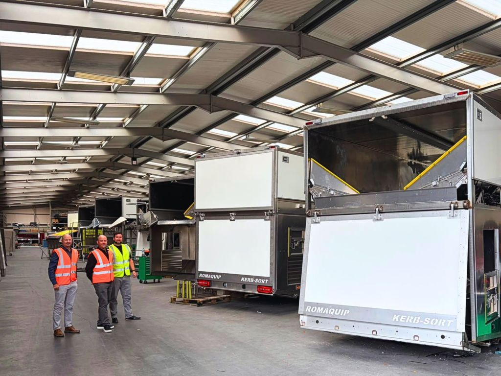 Several recycling collection vehicle bodies lined up inside a large workshop, with three council staff in hi-vis standing on the left.