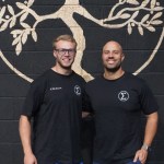 Elysium Training Club co-founders Peter Crudge and Andrew Finnegan stand smiling in front of a tree-of-life mural at their new Swansea gym.