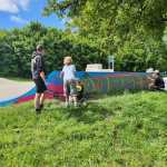 Children and an adult helper spray-painting the "Mumbles" mural on a skatepark ramp, with the artist working at the far end.