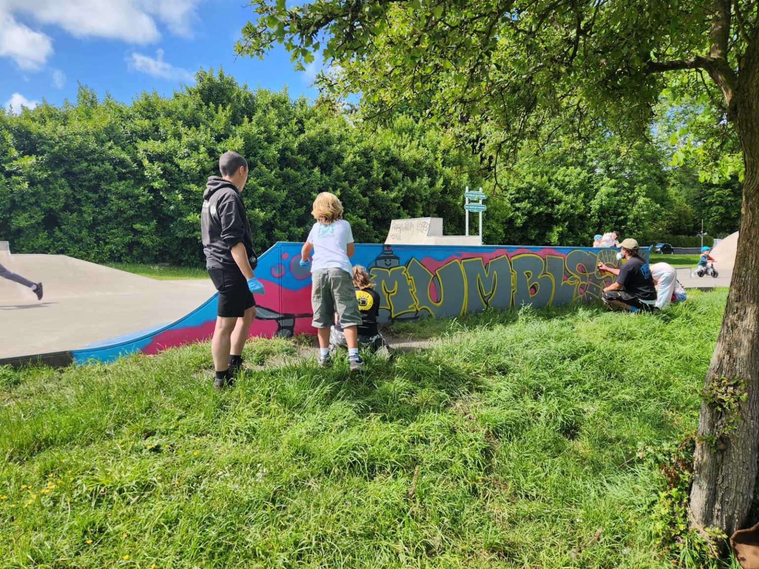 Children and an adult helper spray-painting the "Mumbles" mural on a skatepark ramp, with the artist working at the far end.