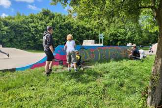 Children and an adult helper spray-painting the "Mumbles" mural on a skatepark ramp, with the artist working at the far end.