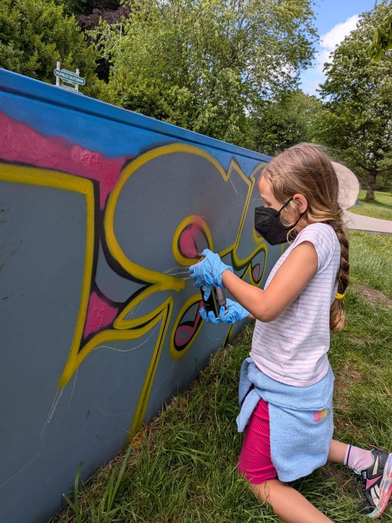 A masked young participant in gloves spray-painting a mural at Mumbles skatepark.